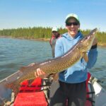 What a catch Katie! Person holding a large fish on a boat with another person and a forested shoreline in the background.