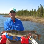 Nice catch John! Man in a blue shirt and green cap holding a large fish on a boat, with water and trees in the background.