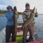 What a double! Two people in a boat hold large fish, standing side by side on a lake with trees in the background.