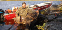 A person in camouflage poses with a large moose by a lake, next to a red boat on the rocky shore.