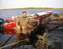 A person in camouflage poses with a large moose by a lake, next to a red boat on the rocky shore.