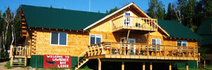 A wooden lodge with a green roof, deck, and a “Welcome to Lawrence Bay Lodge” sign, set on green grass under a blue sky.