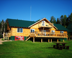 A wooden lodge with a green roof, deck, and a “Welcome to Lawrence Bay Lodge” sign, set on green grass under a blue sky.