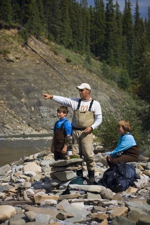 An adult and two children in fishing gear stand and sit on rocks by a river, with forested hills in the background.