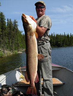 Man standing in a boat on a lake holding a large fish upright with trees visible in the background.