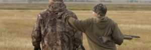 Two people in camouflage jackets walk through a field; one carries a shotgun and points at something in the distance.