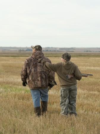 Two people in camouflage jackets walk through a field; one carries a shotgun and points at something in the distance.
