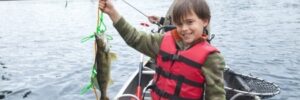 A boy in a red life jacket holds up a fish in a boat on a lake, with an adult fishing behind him.