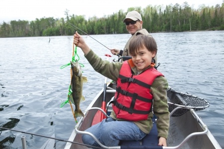 A boy in a red life jacket holds up a fish in a boat on a lake, with an adult fishing behind him.