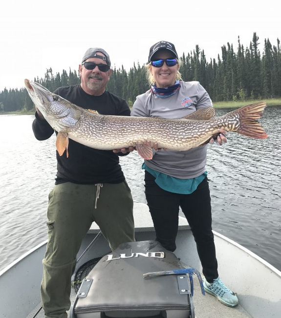 Two people on a boat hold a large fish together, with a lake and forest in the background.