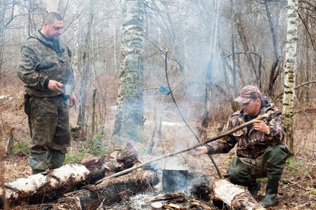 Two men in camouflage clothing stand by a smoky campfire in a forest, one stirring a pot while the other holds a mug.