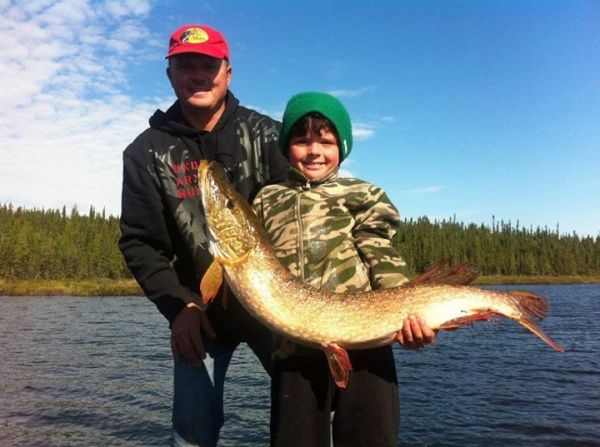 An adult and child pose on a lake holding a large fish, with trees and blue sky in the background.