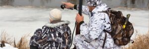 Two people in camouflage winter gear by a snowy riverbank, one pointing across the water, the other holding a shotgun.