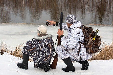 Two people in camouflage winter gear by a snowy riverbank, one pointing across the water, the other holding a shotgun.