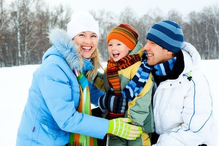 Three people in winter clothing smile outdoors in a snowy landscape with bare trees in the background.