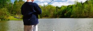 A young boy stands on a wooden dock fishing in a lake, surrounded by trees under a partly cloudy sky.