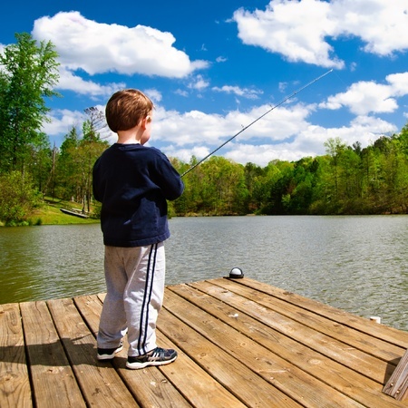 A young boy stands on a wooden dock fishing in a lake, surrounded by trees under a partly cloudy sky.