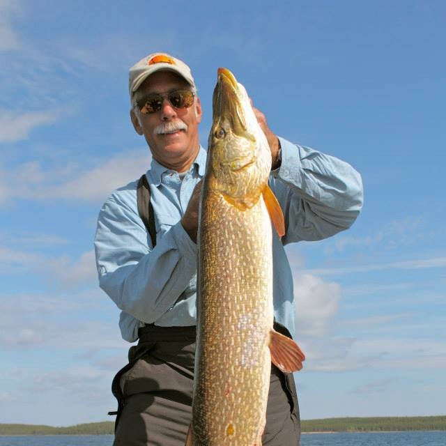 A man in sunglasses holds a large fish outdoors under a blue sky, with water and land visible in the background.