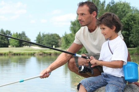 An adult and a child are fishing together by a lakeside, both holding fishing rods and looking at the water.