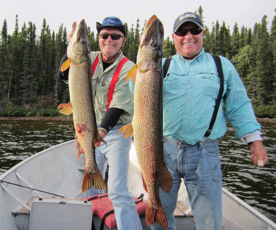 Two men in a boat holding large fish, with forested shoreline in the background.