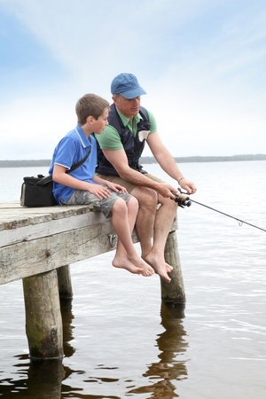 An adult and child sit on a wooden dock fishing, with their feet dangling above the water.