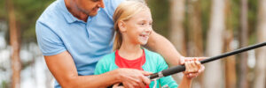 An adult and a child standing outdoors, holding a fishing rod together, with trees in the background.