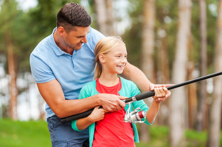 An adult and a child standing outdoors, holding a fishing rod together, with trees in the background.