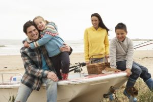 Four people sit on a boat on the beach, smiling, with fishing gear and a picnic basket visible.