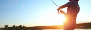 Person in a hat fishing from a boat on a calm lake at sunset, with trees visible in the distance.