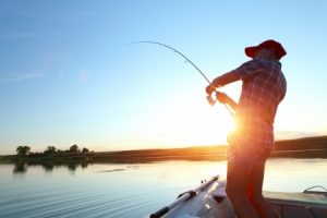 Person in a hat fishing from a boat on a calm lake at sunset, with trees visible in the distance.