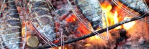 Four whole fish are grilling over an open flame on metal racks above hot coals.