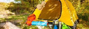 Yellow camping tent set up in a wooded area with backpacks, cookware, and camping gear outside on the grass.