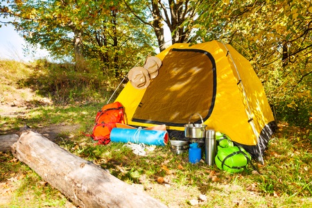 Yellow camping tent set up in a wooded area with backpacks, cookware, and camping gear outside on the grass.