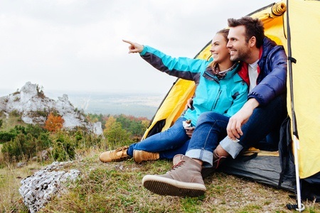 Two people sit at the entrance of a yellow tent on a hillside, looking and pointing at the landscape ahead.