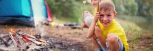 Two children roast marshmallows over a campfire near a blue tent in a wooded outdoor setting.