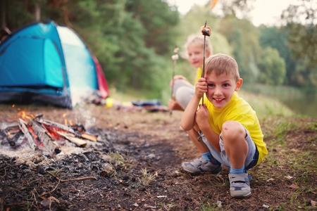 Two children roast marshmallows over a campfire near a blue tent in a wooded outdoor setting.