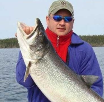 A person wearing sunglasses and a cap holds a large fish on a boat with water and trees in the background.