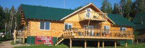 Log cabin-style lodge with green roof, wooden deck, and a "Welcome to Lawrence Bay Lodge" sign out front.