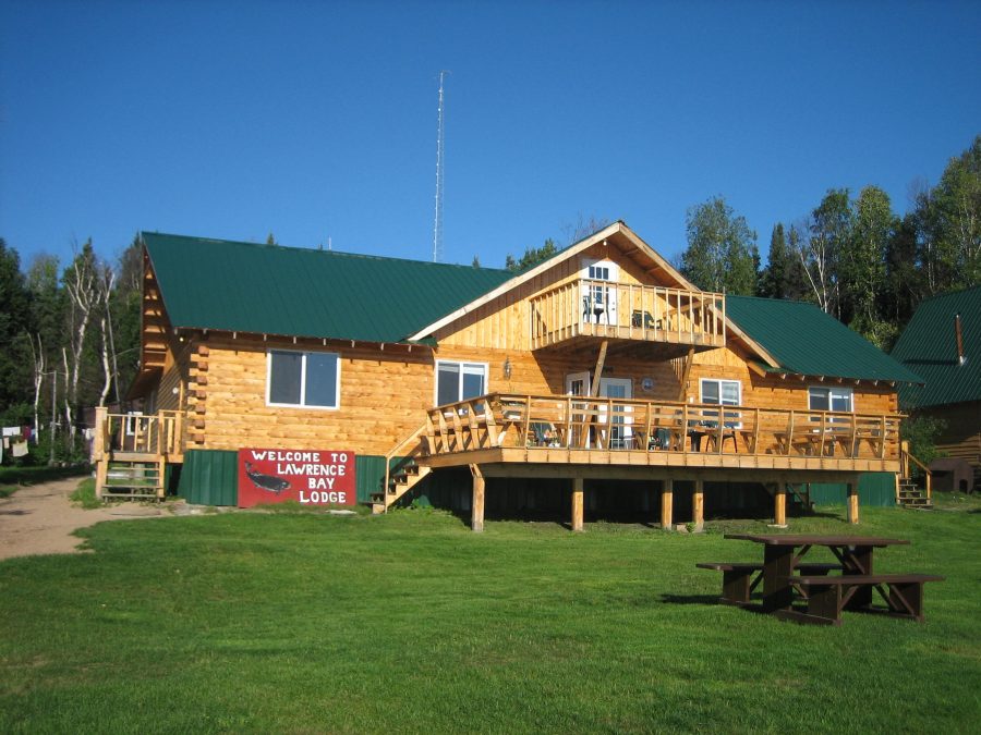 Log cabin-style lodge with green roof, wooden deck, and a "Welcome to Lawrence Bay Lodge" sign out front.