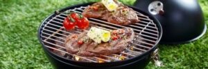 Two seasoned steaks and tomatoes grilling on a small charcoal barbecue, placed on green grass.