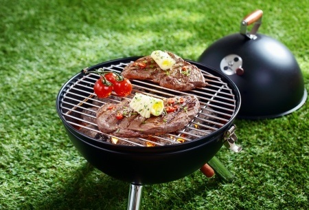 Two seasoned steaks and tomatoes grilling on a small charcoal barbecue, placed on green grass.