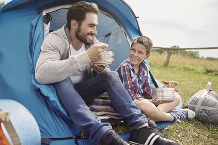 An adult and a child sit outside a blue tent, eating food and smiling at each other.