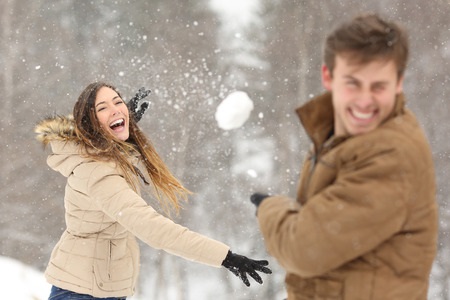 A woman throws a snowball at a man outdoors in a snowy setting, both wearing winter coats and gloves.