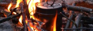 A metal kettle heating over a campfire with burning sticks and flames visible.
