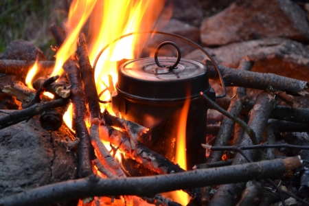 A metal kettle heating over a campfire with burning sticks and flames visible.