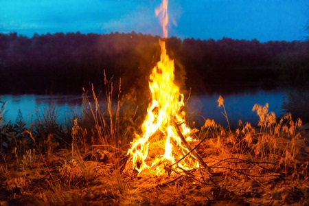 A campfire burns brightly near a lake at dusk, surrounded by dry grass and trees in the background.