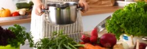 Person in striped apron holding a pot in a kitchen with various fresh vegetables on the counter.