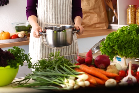 Person in striped apron holding a pot in a kitchen with various fresh vegetables on the counter.