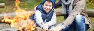 An adult and child roast marshmallows over a campfire outdoors, both smiling and wearing warm clothing.