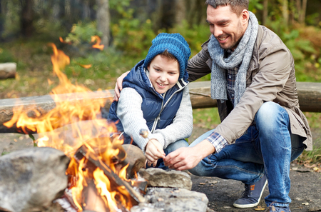 An adult and child roast marshmallows over a campfire outdoors, both smiling and wearing warm clothing.